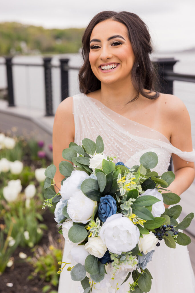 Bride smiles while holding bouquet in front of spring florals on the terrace at Water’s Edge Centerport.