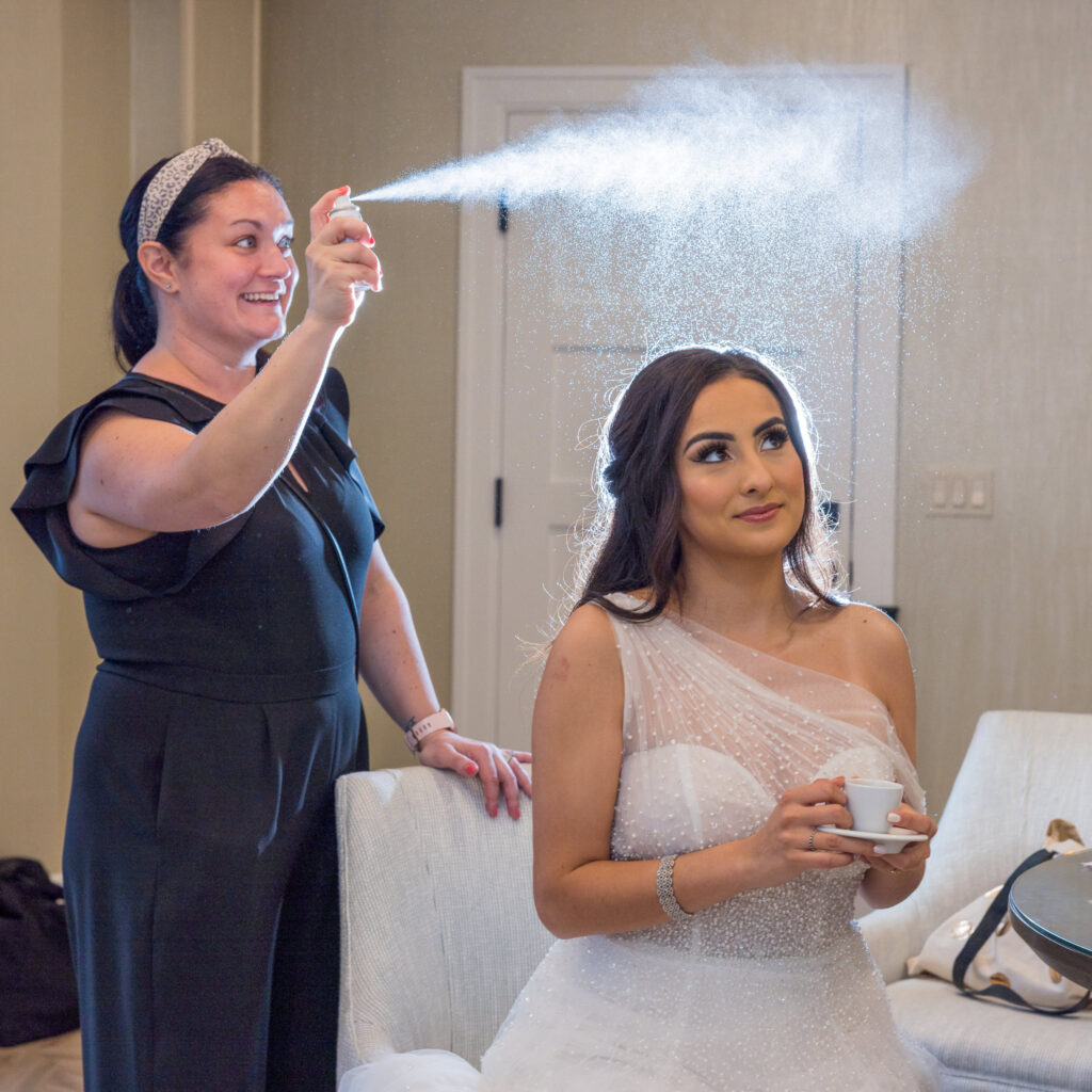 Bride sits calmly as hairstylist applies finishing spray during wedding prep at Water’s Edge Centerport.