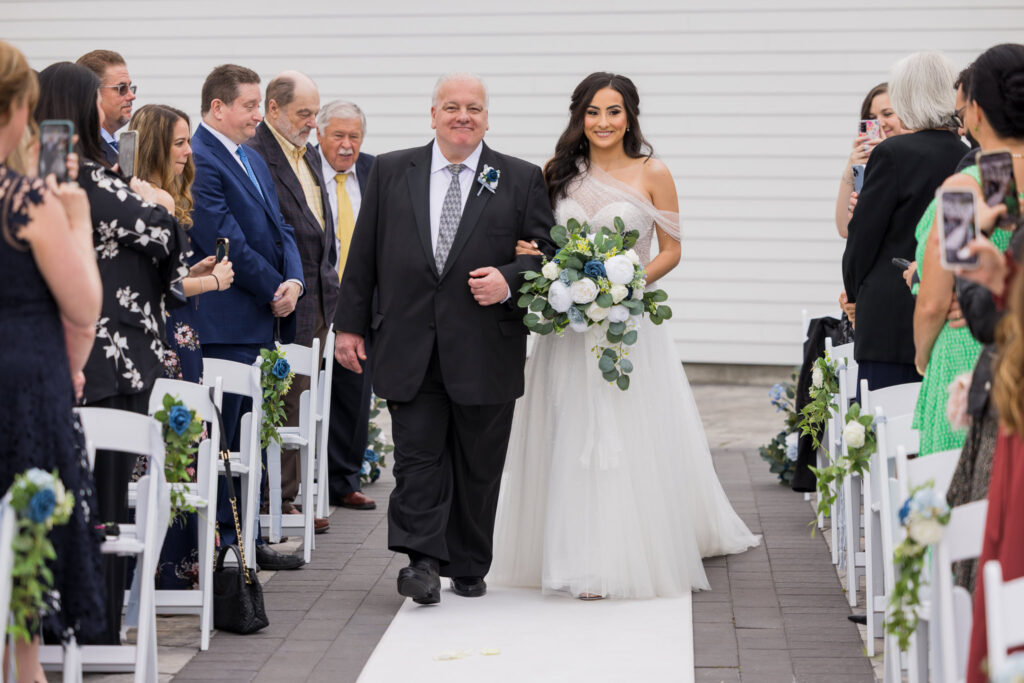 Bride walks down the aisle arm-in-arm with her father, surrounded by guests at Water’s Edge Centerport.