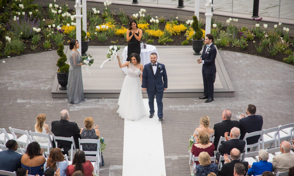 Bride and groom raise their hands in celebration as they exit the ceremony at Water’s Edge Centerport.