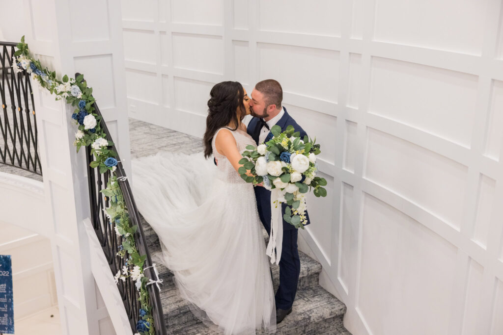 Bride and groom share a kiss on the staircase holding a lush white and blue bouquet at Water’s Edge Centerport.