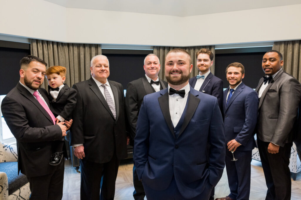 Groom stands proudly with his groomsmen and family in the getting-ready suite at Water’s Edge Centerport.