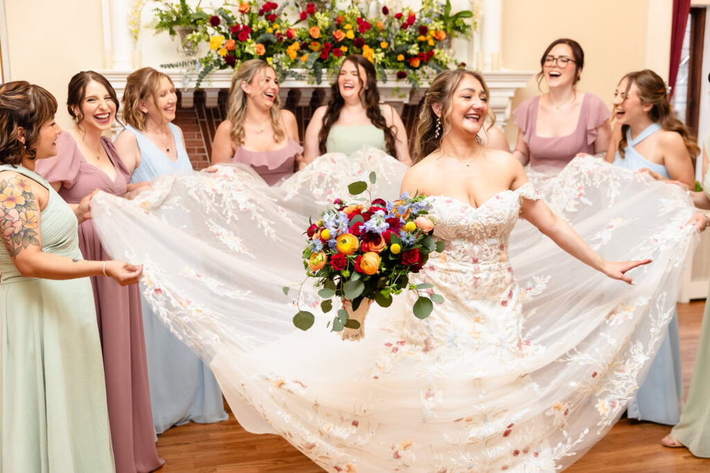 Bride beaming with joy as bridesmaids hold her gown in bridal suite