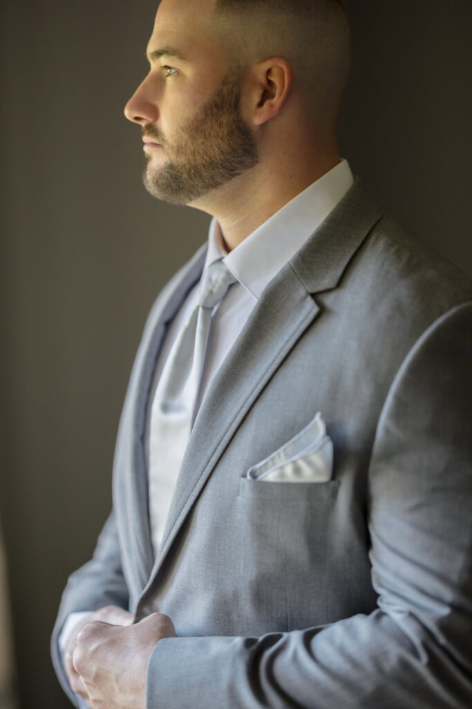 Groom in a light gray suit adjusting his jacket, softly lit from the side as he prepares for the wedding day.

