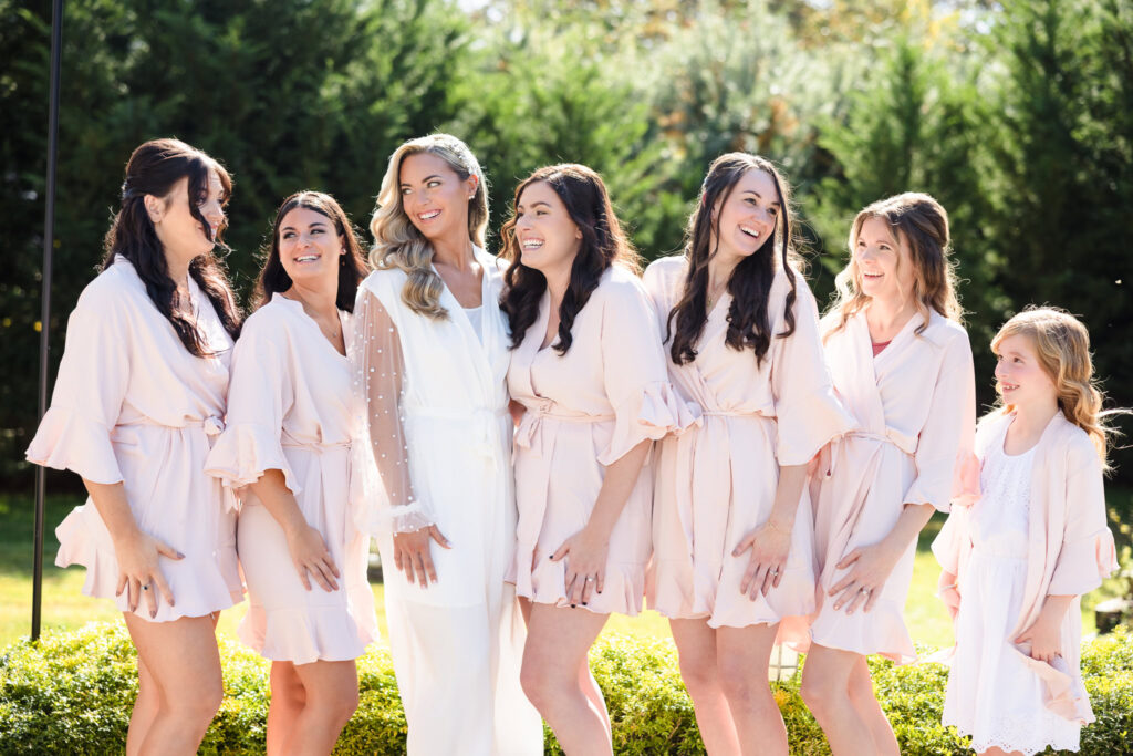 Bride laughing with her bridesmaids and flower girl in soft blush robes during a sunlit morning getting ready session.

