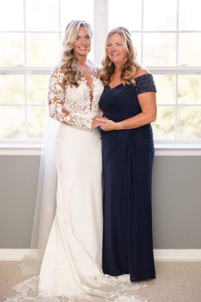 Bride standing beside her mother by a sunlit window, both smiling and holding hands in an intimate pre-ceremony portrait.

