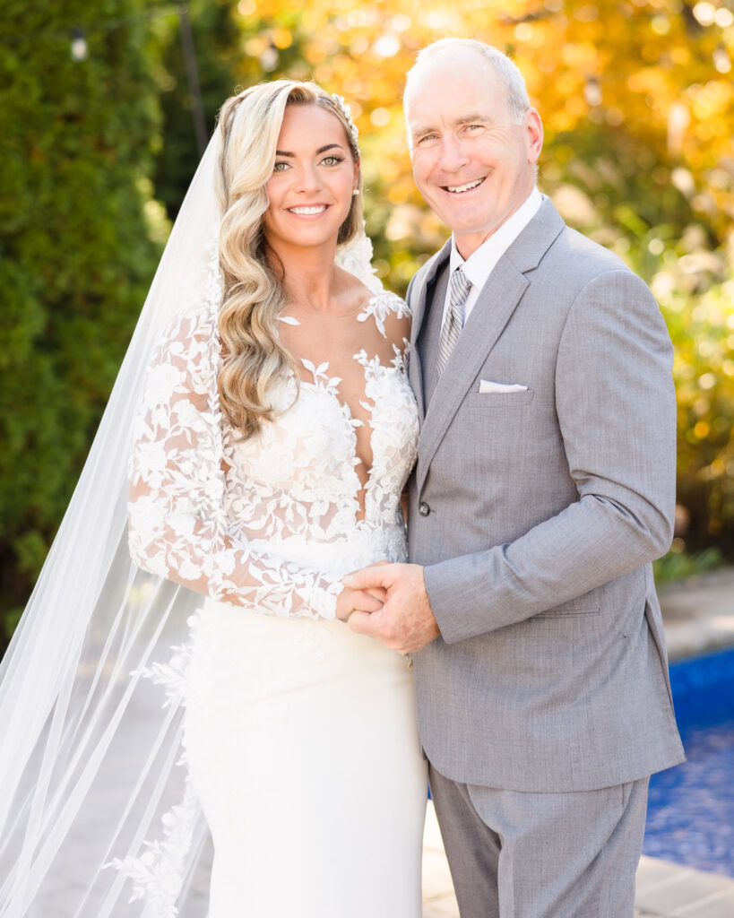 Bride smiling with her father on her wedding day, standing outdoors with vibrant fall foliage behind them.

