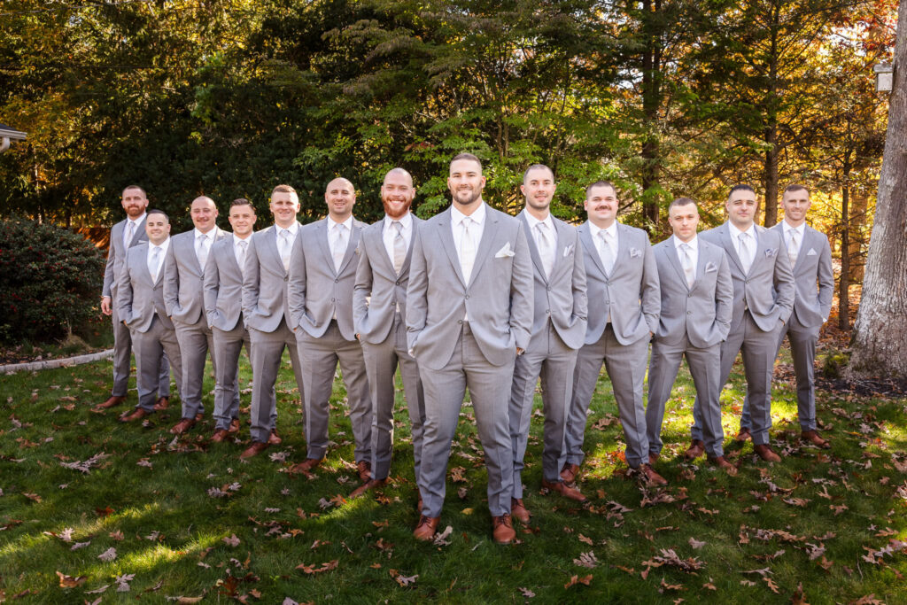 Groom and groomsmen in light gray suits standing in formation under dappled autumn light, ready for the ceremony.

