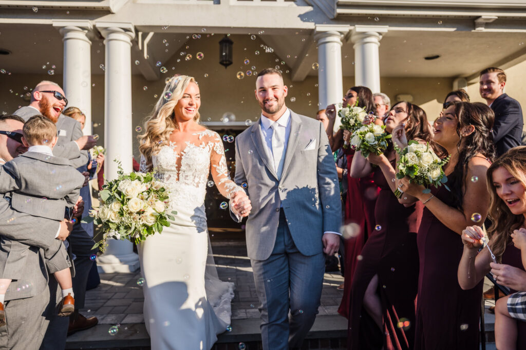 Just-married couple exits the church hand in hand as guests blow bubbles and cheer in the afternoon sun.
