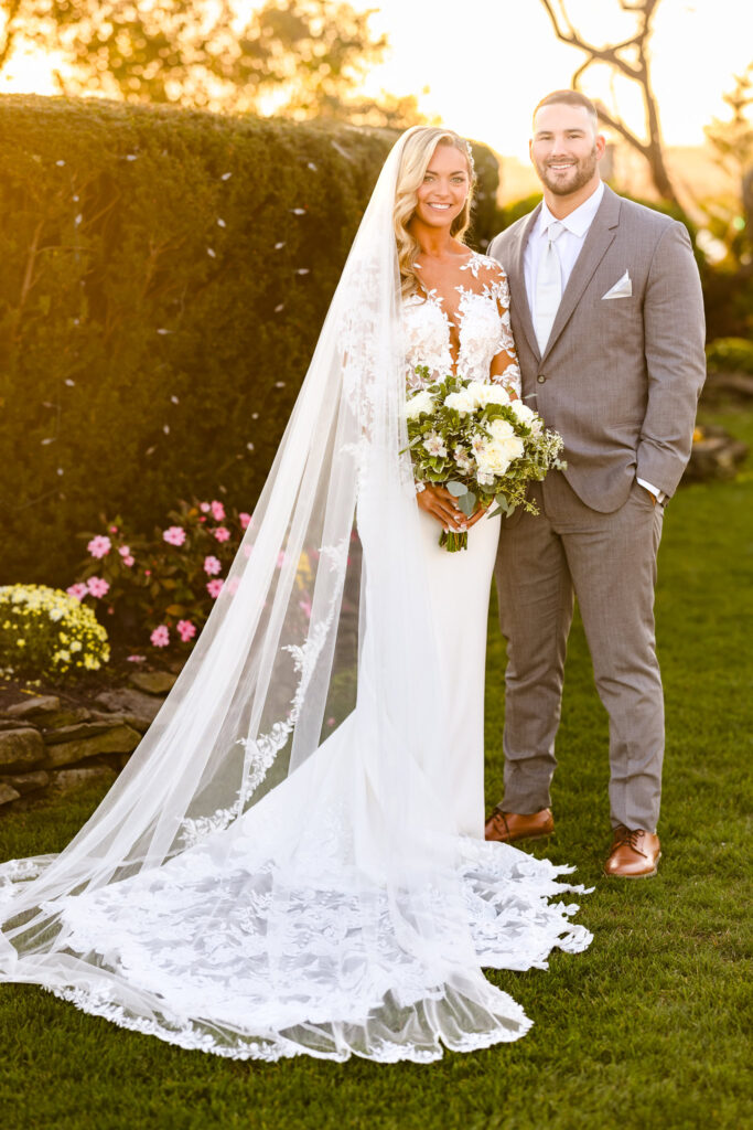 Bride and groom standing together in a sunlit garden at Land’s End, with her lace veil draped behind and golden light surrounding them.

