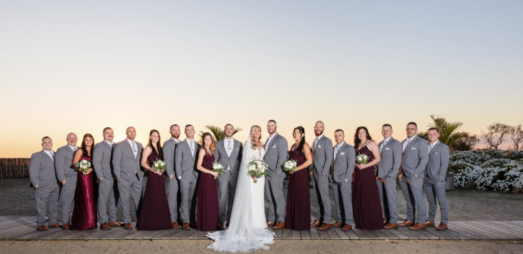 Full wedding party standing together on the boardwalk at Land’s End during sunset, with the bride and groom centered and the bridal party in slate gray and deep plum attire.

