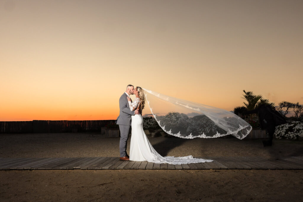 Bride and groom share an intimate embrace on the boardwalk at sunset, with her long lace veil caught in the wind against a glowing sky.

