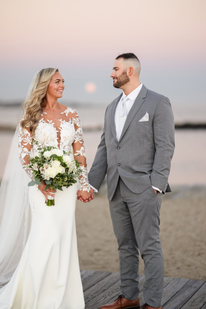 Bride and groom holding hands on the boardwalk at sunset, with a soft pastel sky and the moon rising over the bay in the background.

