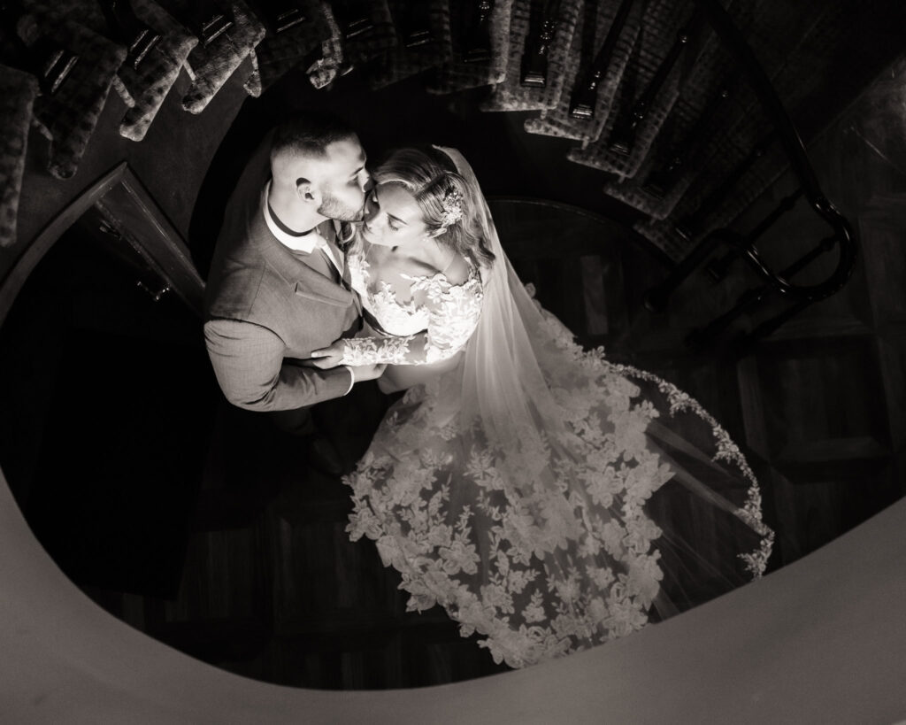Black and white photo of the bride and groom embracing on a spiral staircase, her lace train elegantly fanned out below.

