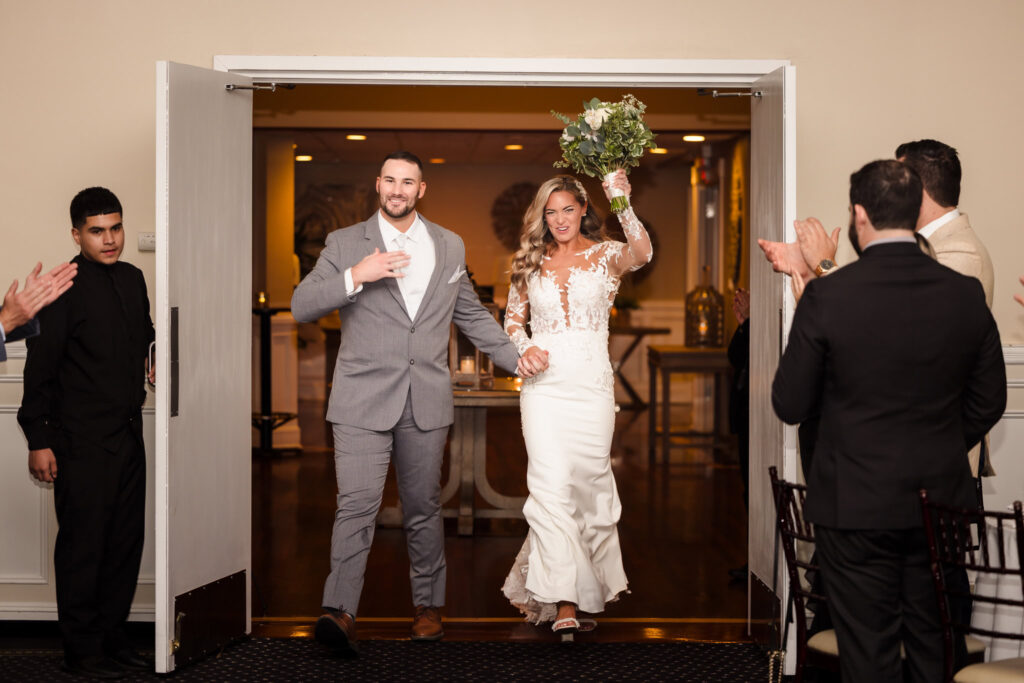Bride and groom make their grand entrance into the reception, hand in hand and smiling as guests cheer and applaud.

