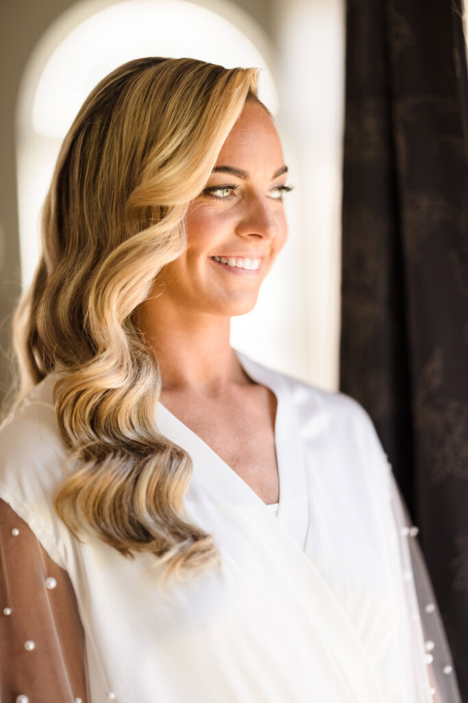 Bride standing by the window in a soft white robe, glowing in natural light with soft Hollywood waves and a radiant smile.

