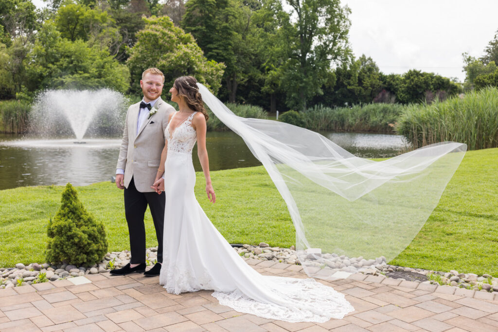 Bride and groom share a quiet moment by the pond at Flowerfield, her long veil flowing in the breeze as a fountain gently sprays in the background.

