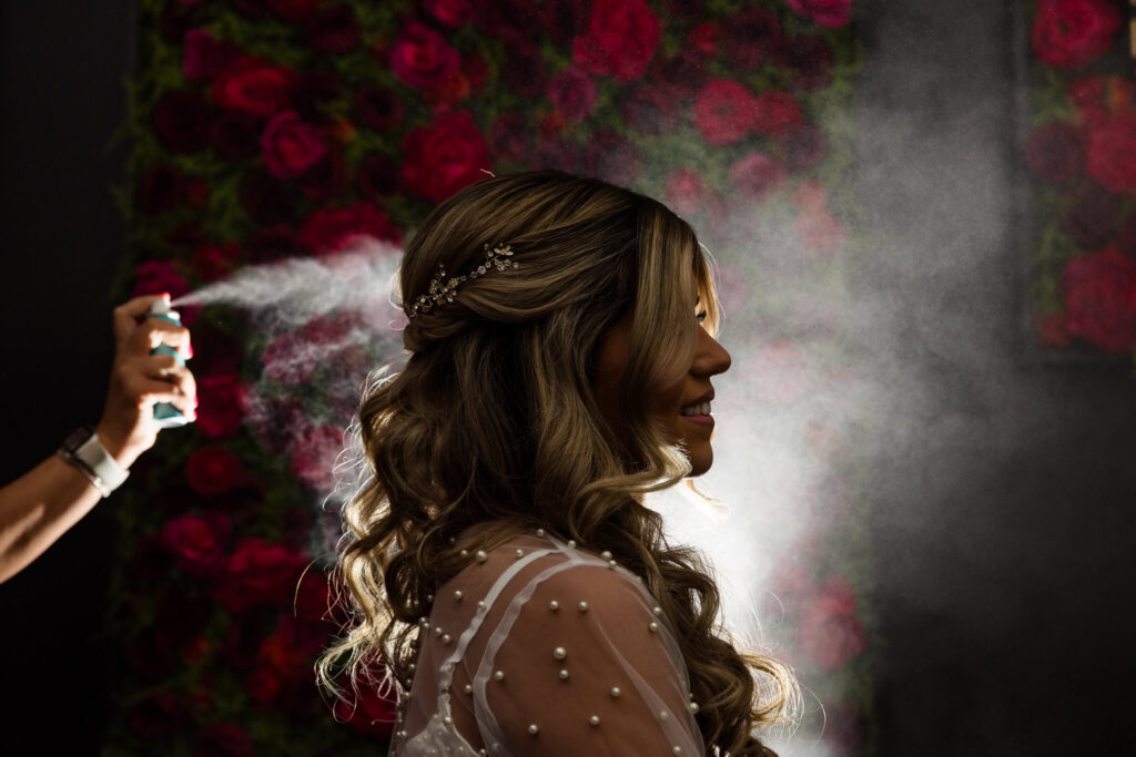 A soft mist catches the light as the bride’s stylist sets her curls, silhouetted against a floral backdrop in a serene, cinematic getting-ready moment.

