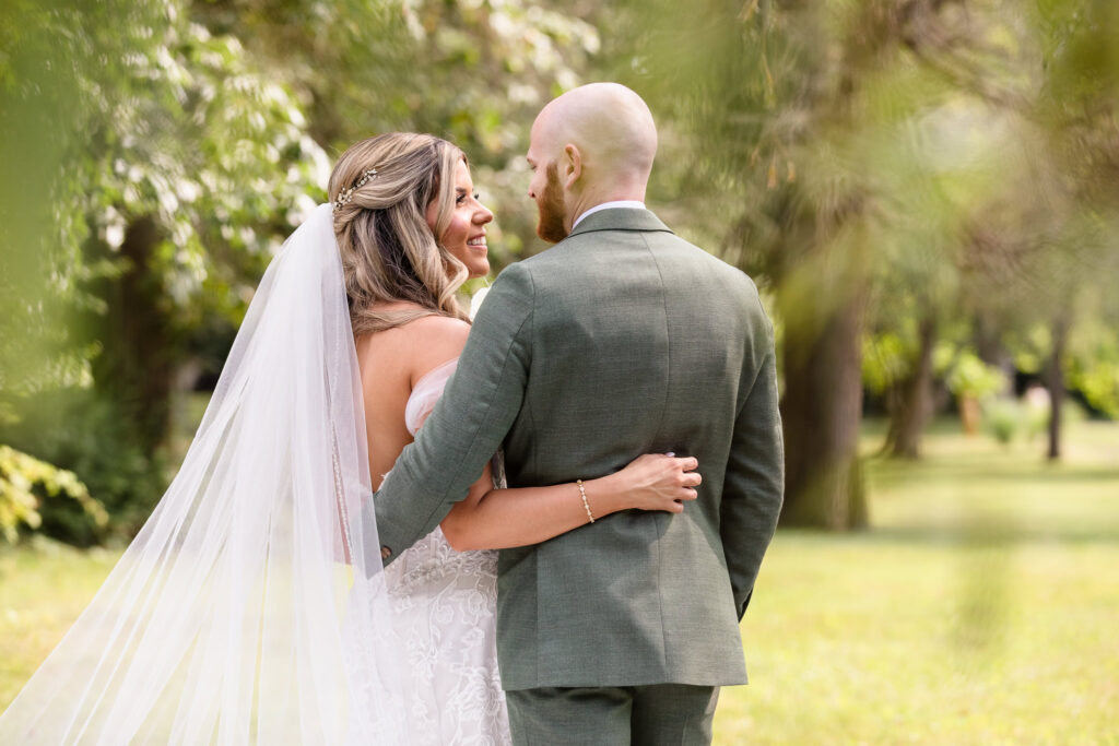 Bride and groom embrace in a quiet moment on the lush grounds of Flowerfield, smiling at one another beneath soft afternoon light and surrounded by nature.