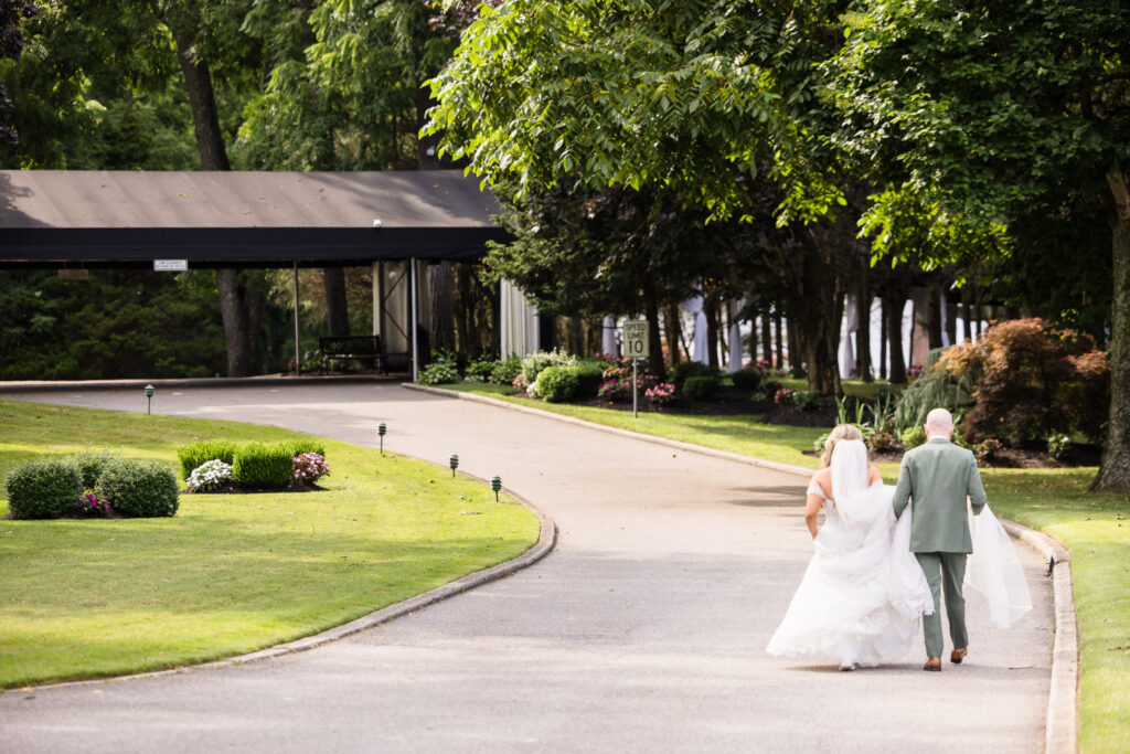 Bride and groom walk hand-in-hand down the winding driveway at Flowerfield, her gown gently lifted as they stroll beneath the afternoon light toward the covered entrance.

