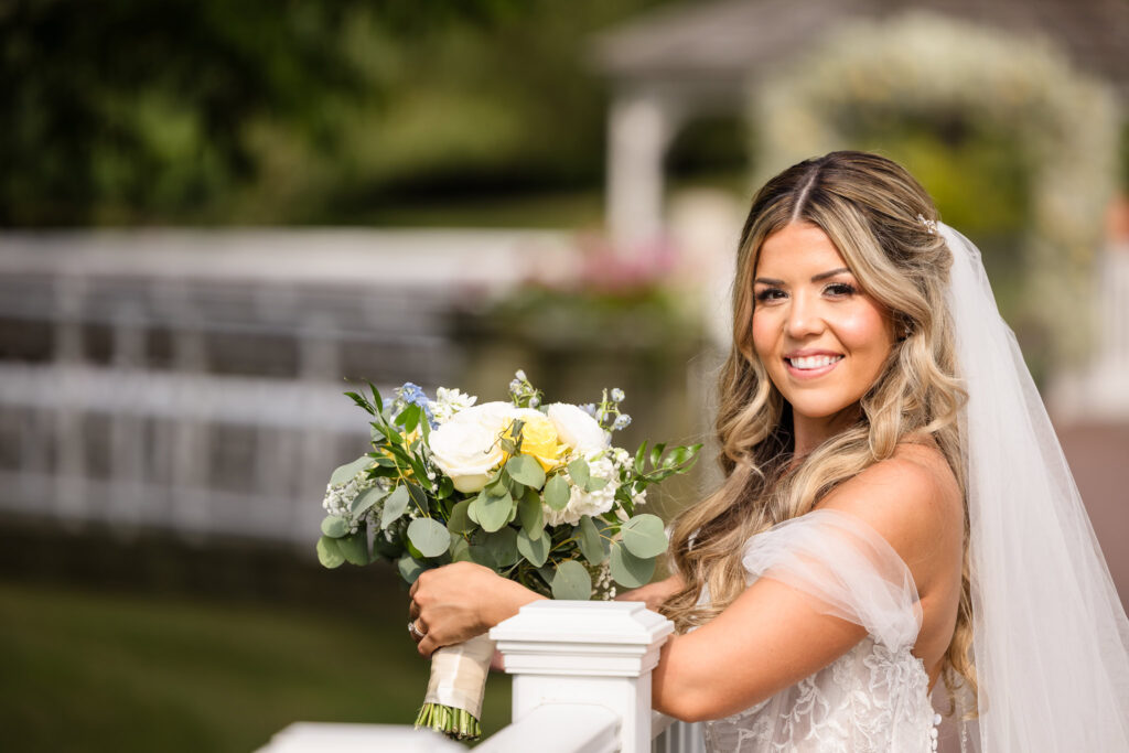 Bride smiles softly while holding her bouquet along the white bridge at Flowerfield, her veil flowing and sunlight casting a golden glow on her timeless, romantic look.

