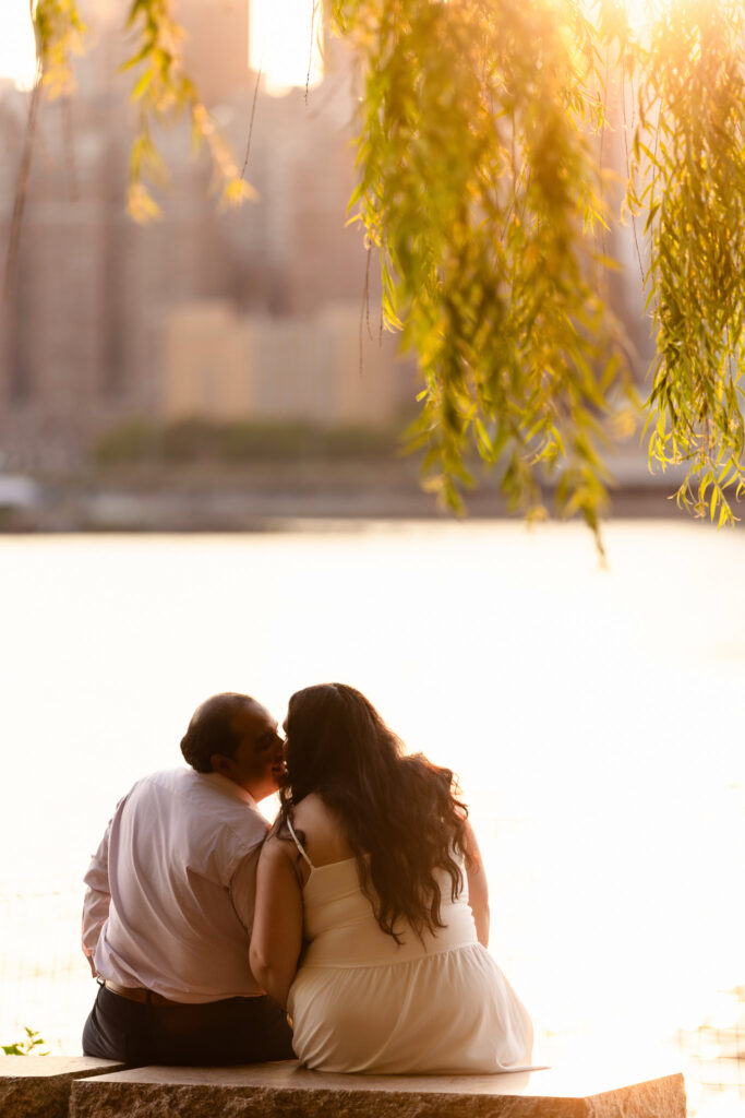 Couple sitting close together by the East River, framed by soft golden light and willow branches at Gantry Plaza State Park.
