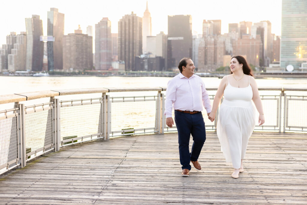 Couple walking hand in hand along the Gantry Plaza boardwalk with Manhattan’s skyline softly glowing behind them.