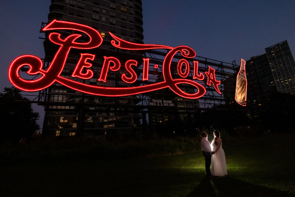 Couple standing beneath the iconic Pepsi-Cola sign with city lights twinkling in the background.