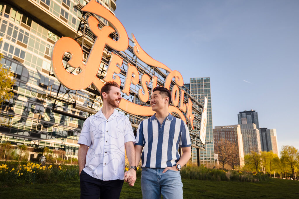 Dave and Peter walk hand in hand beneath the iconic Pepsi-Cola sign in Long Island City, framed by spring skies and city energy.
