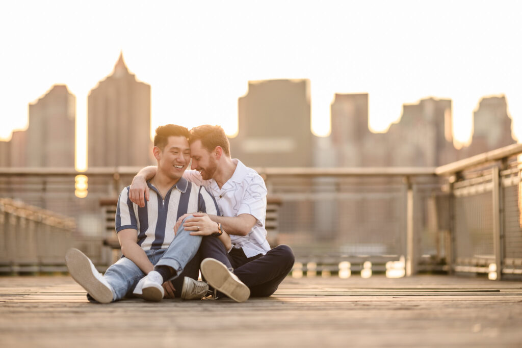 Seated on the boardwalk with the skyline behind them, the couple leans in with laughter and lightness, surrounded by evening calm.