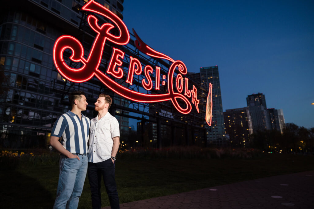 Dave and Peter stand beneath the glowing red Pepsi-Cola sign, with twilight settling over the Long Island City skyline.