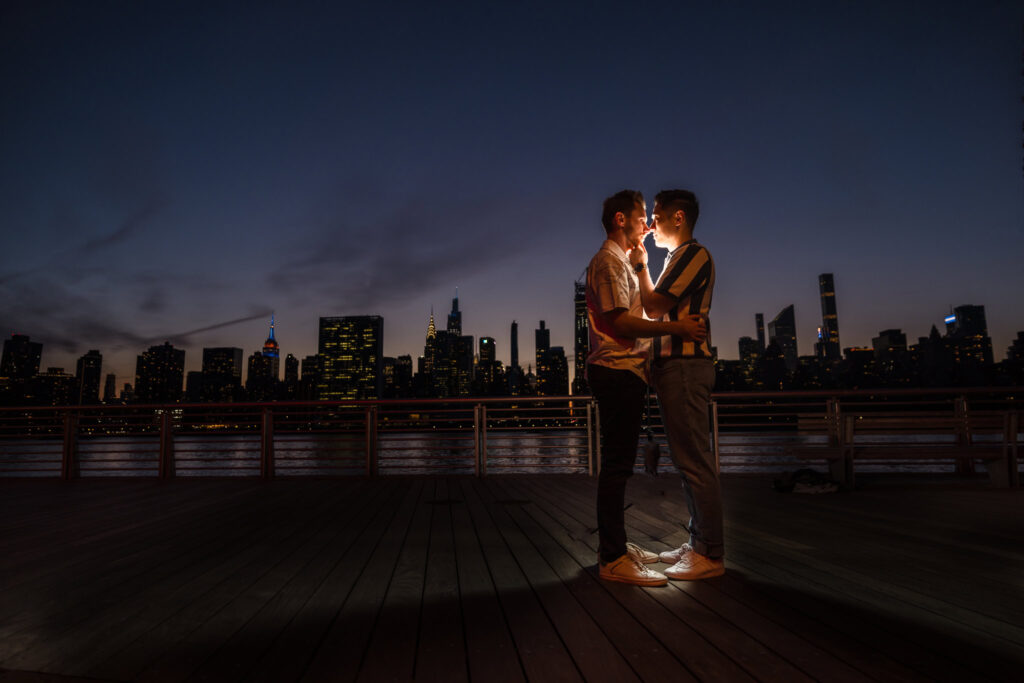 The couple embraces on the boardwalk at night, softly lit against the glowing Manhattan skyline, full of emotion and warmth.