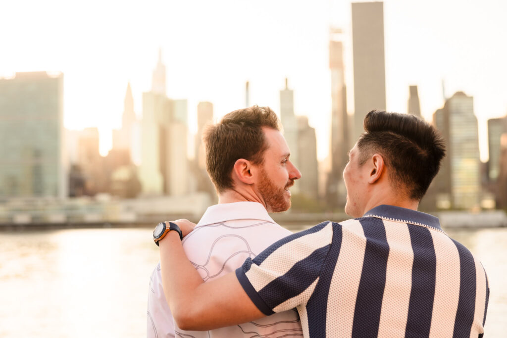 Dave and Peter turn toward one another, smiling as warm sunset light reflects off the East River behind them.