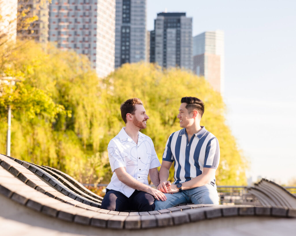 Seated together in Gantry Plaza, Dave and Peter share a lighthearted exchange surrounded by spring foliage and city textures.