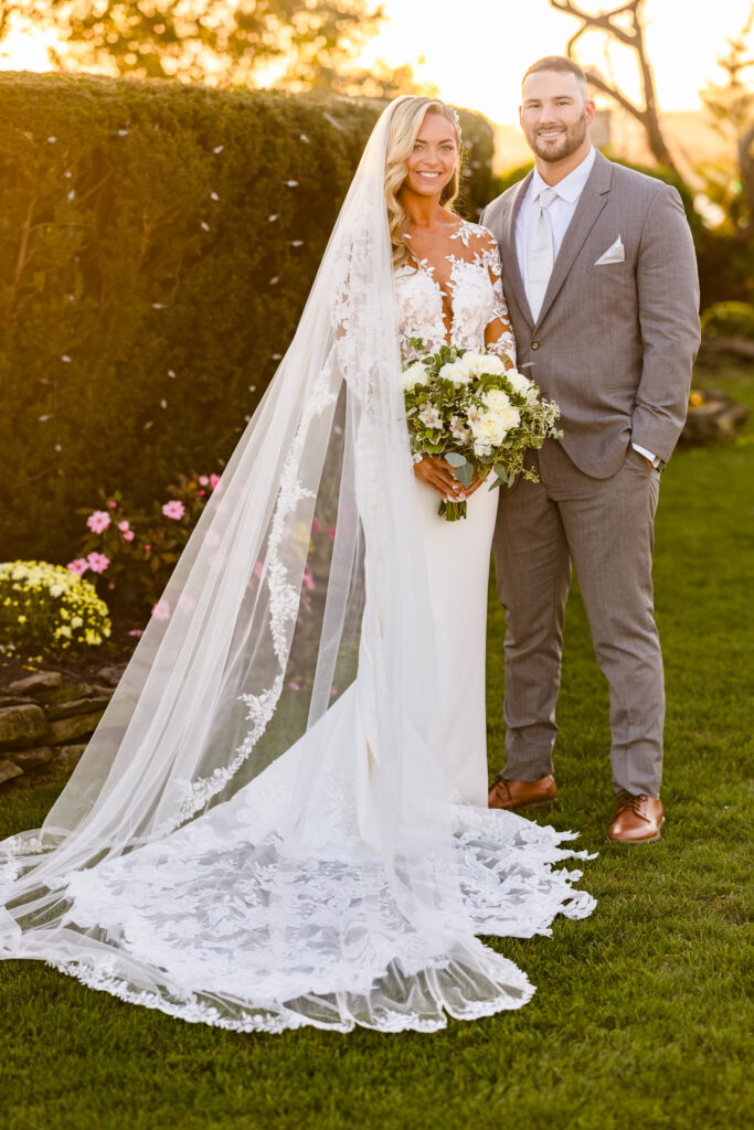 Bride and groom standing together in a sunlit garden at Land’s End, with the bride in a long lace-trimmed veil holding a lush bouquet.

