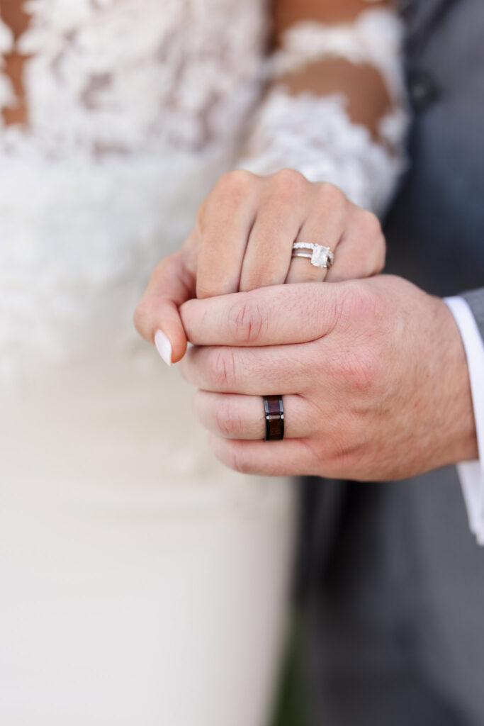 Close-up of the couple’s hands showing their wedding rings, with the bride’s lace sleeve and gown softly in focus.

