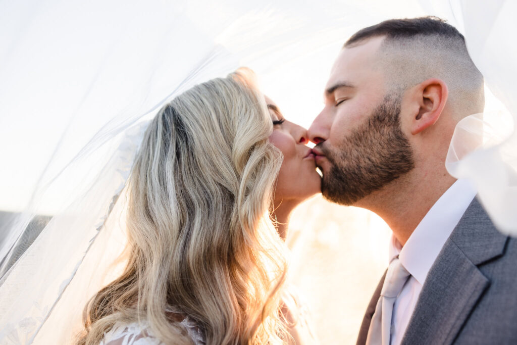 Close-up of bride and groom sharing a kiss under her veil, softly backlit for a romantic and ethereal feel.

