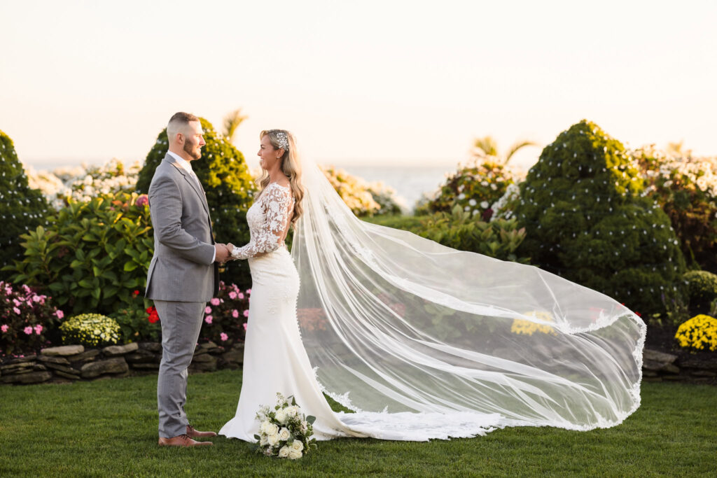 Bride and groom holding hands in a garden at Land’s End, with her cathedral veil flowing behind and the bay softly visible in the distance.

