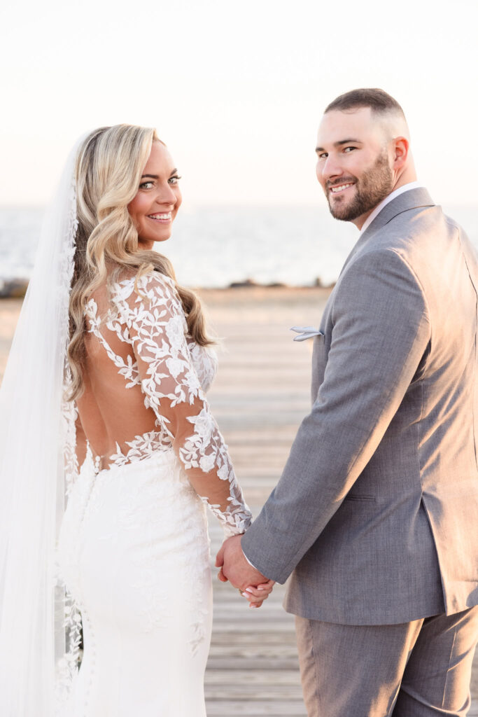 Bride and groom holding hands on the boardwalk at Land’s End, smiling back toward the camera with the bay behind them.


