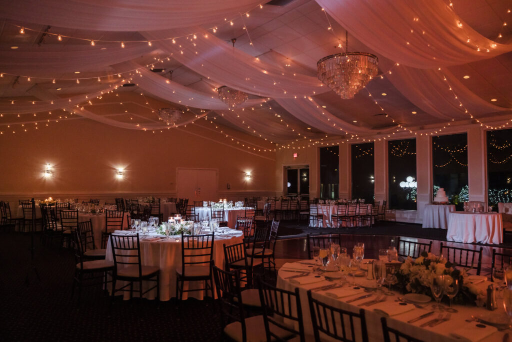 The reception ballroom at Land’s End set for evening, with draped ceilings, chandeliers, and string lights creating a warm, elegant glow.

