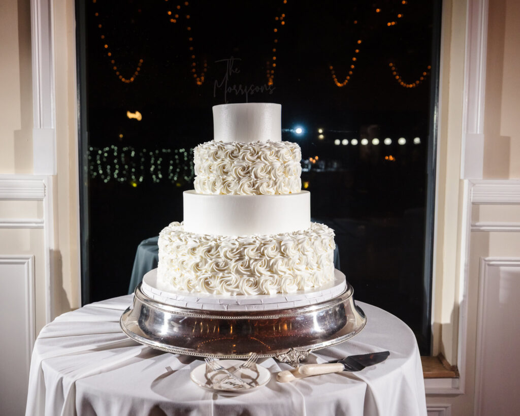 Three-tier white wedding cake with rosette detailing and custom topper, displayed by the window at Land’s End with twinkle lights in the background.

