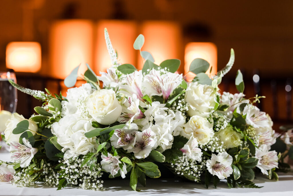 Close-up of a white and blush floral centerpiece with roses, lilies, eucalyptus, and baby's breath, softly lit against a warm reception backdrop.

