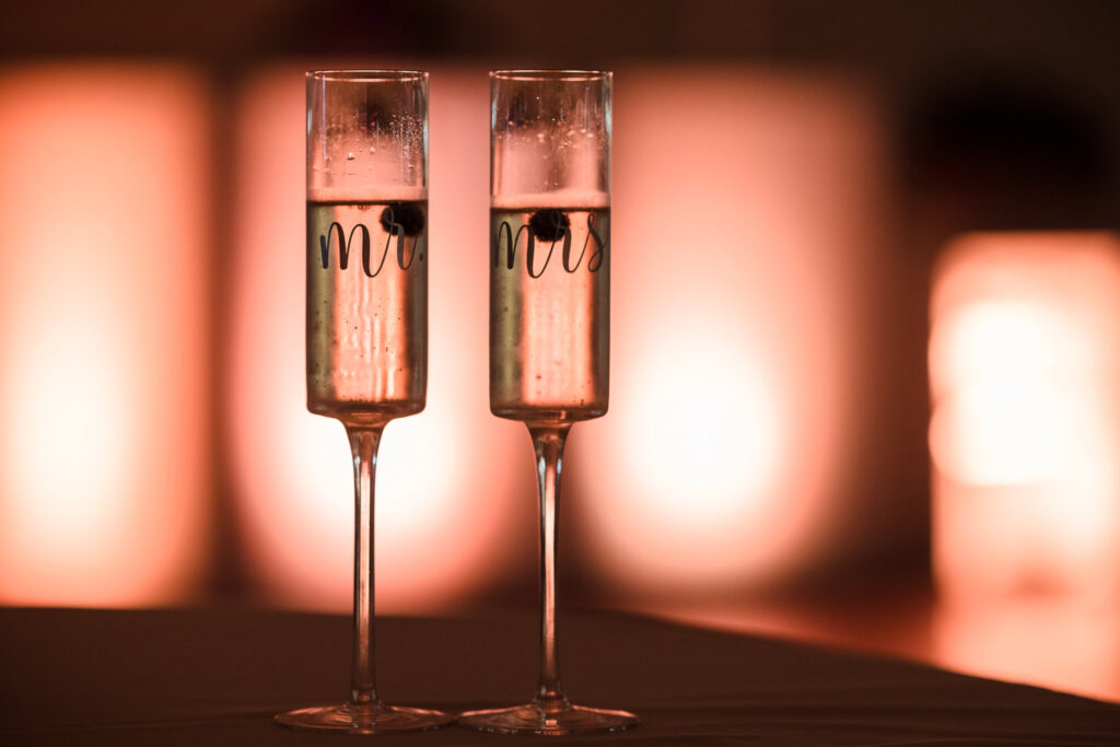 Close-up of two champagne flutes labeled “Mr.” and “Mrs.” filled with bubbly, softly backlit for a romantic evening ambiance.

