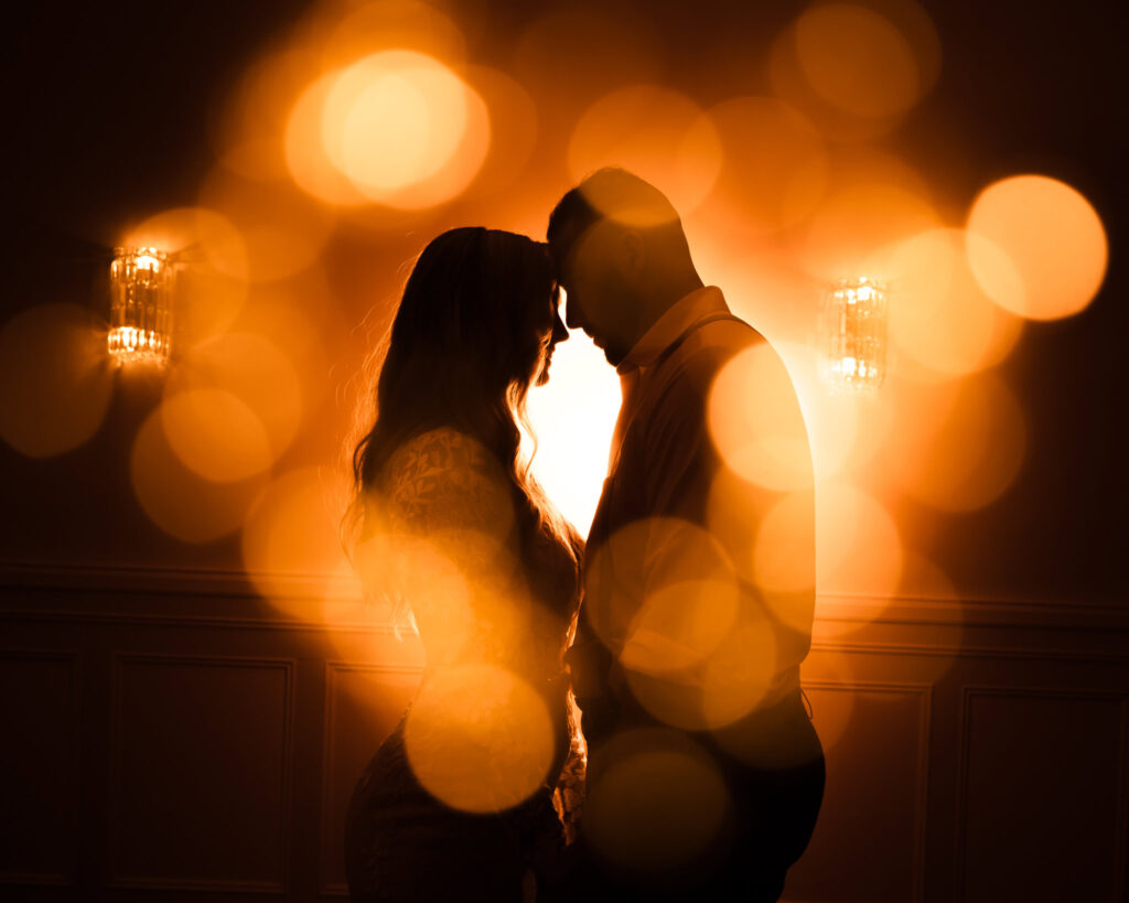 Silhouette of a bride and groom touching foreheads, surrounded by glowing amber bokeh light for a romantic and cinematic effect.