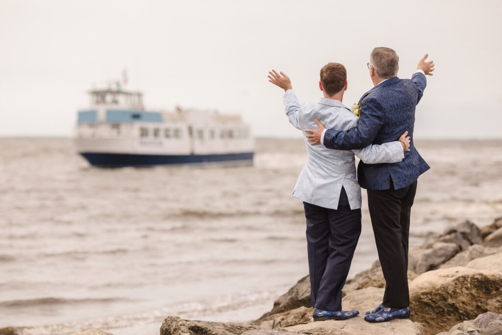 Two grooms standing arm-in-arm on the rocks at Land’s End, joyfully waving toward a passing boat on the bay.