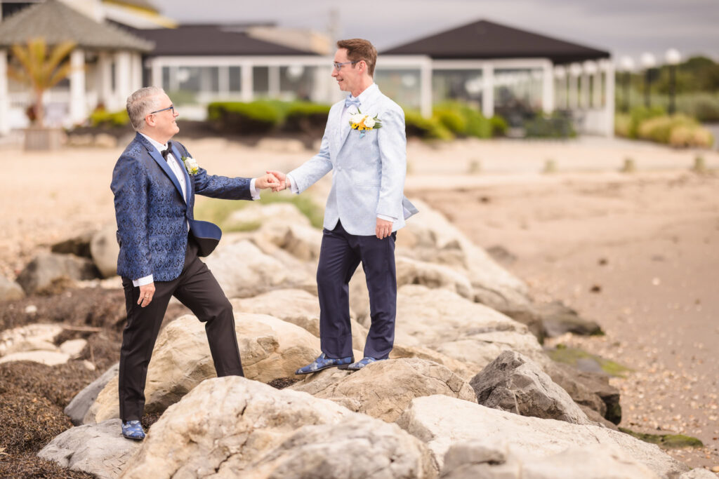 Two grooms holding hands on the rocky shoreline at Land’s End, smiling at each other in tailored blue jackets with boutonnières.

