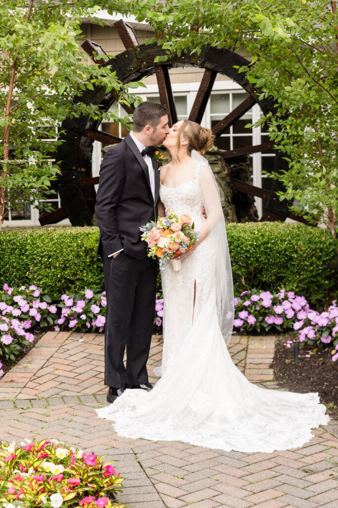 Newlyweds kissing in front of the iconic waterwheel surrounded by gardens at the Watermill wedding venue on Long Island.