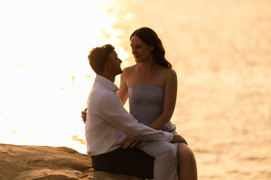 Bride sitting on groom’s lap with golden water in the background