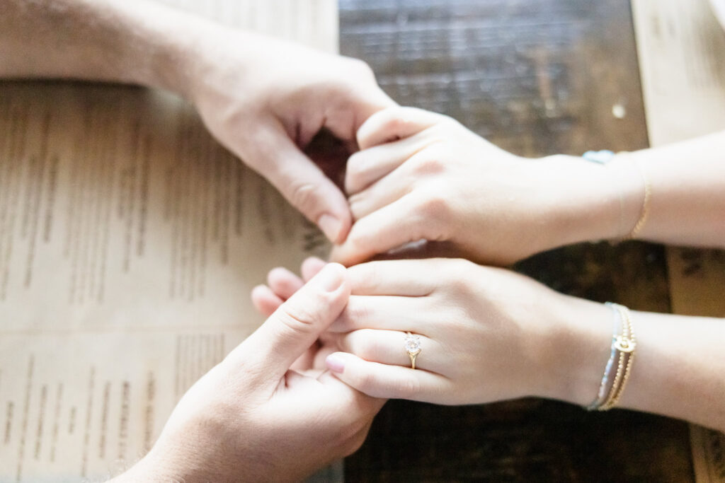 Close-up of their hands intertwined on a wooden table, engagement ring visible.