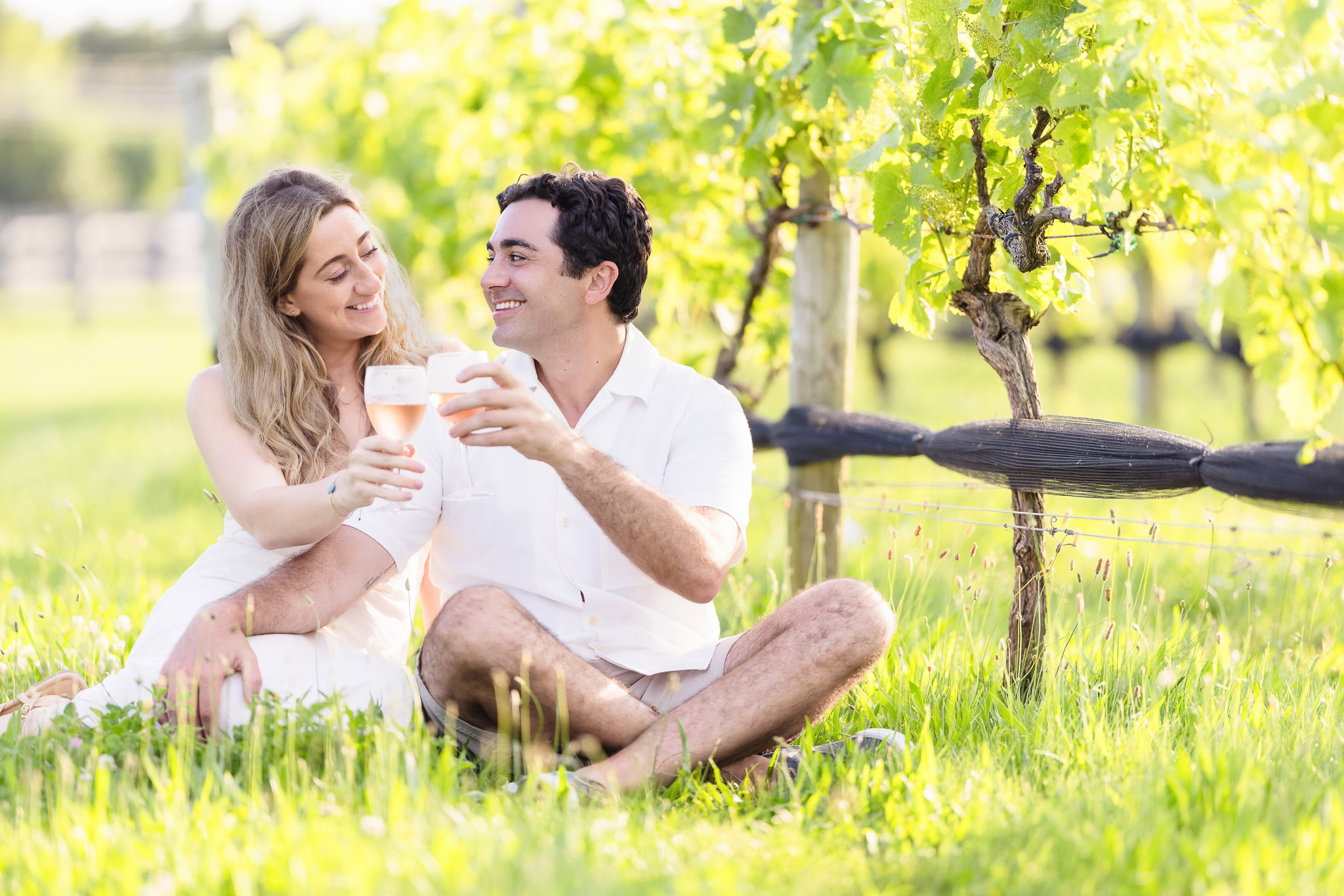 Kristen and James sharing wine in the vineyard, relaxed and joyful on the grass.
